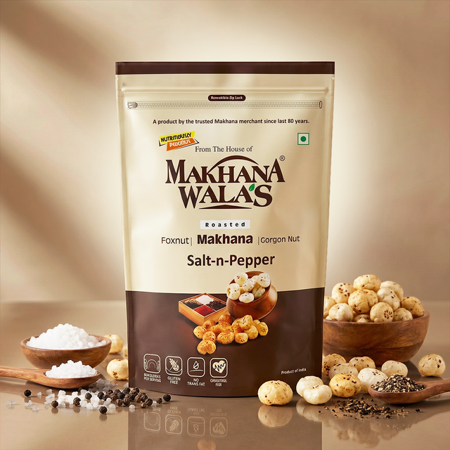A stand-up pouch of Makhana Wala's Roasted Salt-n-Pepper foxnut and corgon nut snack, styled on a reflective surface with bowls of whole and ground spices and salt in the foreground against a warm beige background.