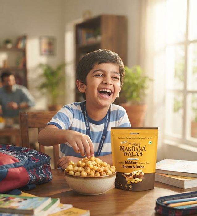 A young boy laughing happily at a wooden table, reaching for popcorn from a bowl. A yellow and brown bag of Makhana Wala's Cream & Onion is prominently displayed next to him, with school supplies like a backpack and pencil case around him in a warm, brightly lit home environment.
